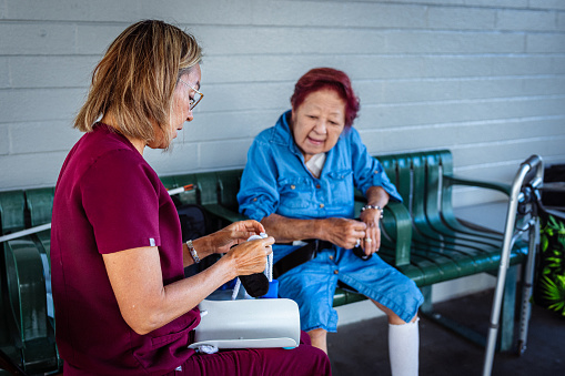 An elderly woman of Asian descent watches as her female occupational therapist teaches her to use a device designed to aid those with limited mobility to put on socks.