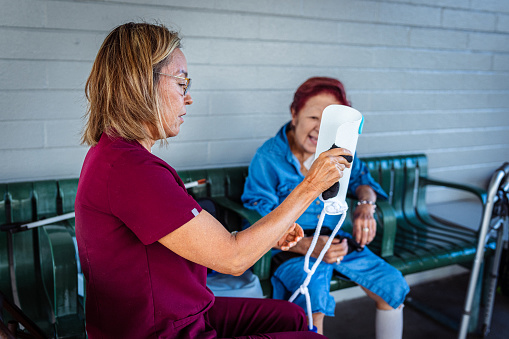 An elderly woman of Asian descent watches as her female occupational therapist teaches her to use a device designed to aid those with limited mobility to put on socks.