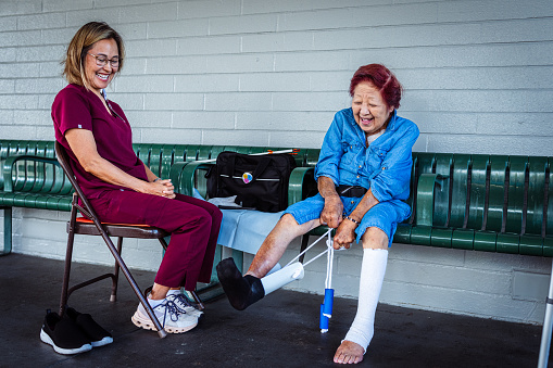 A geriatric woman of Asian descent who has limited mobility sits outside with her occupational therapist and smiles as she practices using a device designed to help her get her shoes and sock on independently.