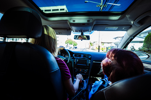 View shot from rear seat of car toward a female home health caregiver driving a vehicle with her elderly patient seated in the passenger seat.