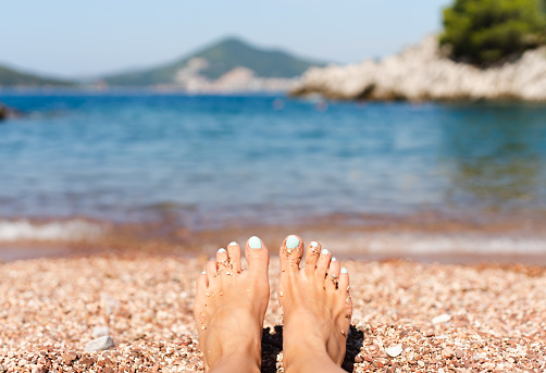 Feet of young woman with pedicure on a pebble beach on vacation. Enjoying the sun on a summer sunny day at the sea. Vacation and travel concept. Close-up.