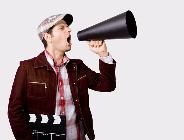 Young man wearing a flat hat, red plaid shirt and maroon jacket shouting on old fashioned megaphone while holding film slate.Megaphone is in his left hand.He is located on the left side of frame and looking at the right side.The photo is taken in studio on white background in horizontal composition.