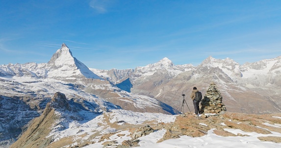 Panoramic landscape of Young man backpacker take a photo by camera at Switzerland mountain peak view point with iconic famous landscape Matterhorn background. Nature, Travel and Adventure concept.