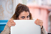 Confident young woman hiding behind laptop computer, looking at camera, spying, peeping.