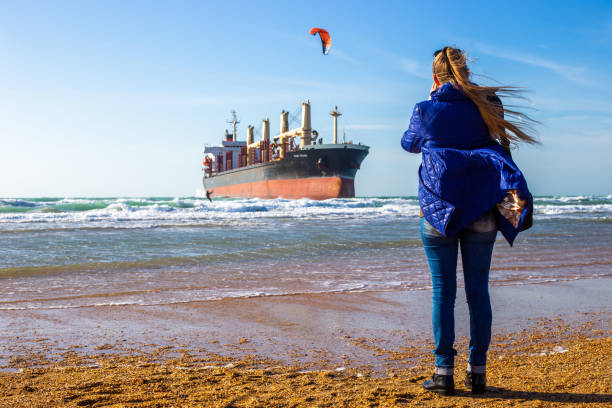 una mujer filma con su teléfono un gran buque de carga en la orilla del mar que ha encallado y un kitesurfista. vityazevo, rusia - stranded men telephone beach fotografías e imágenes de stock