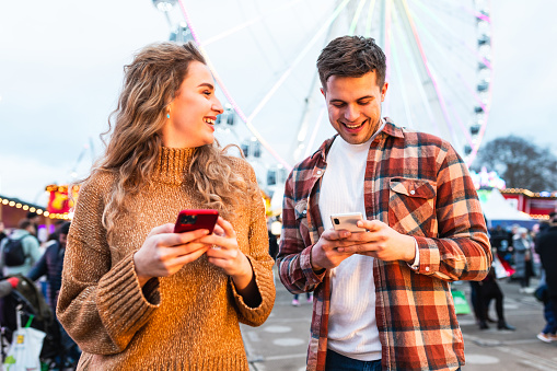Happy couple at amusement park looking at mobile phone and laughing - young couple having fun together on a night out at fun fair - love and happiness concepts