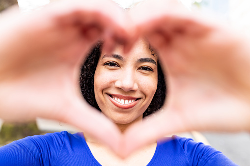 Happy young woman gesturing heart shape with hands, looking through making a nice smile to the camera - Portrait of beautiful black curly woman showing love sign