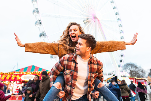 Happy couple having fun at amusement park in London - Portrait of young couple in love enjoying time at funfair with rollercoaster on background - Happy lifestyle and love concepts