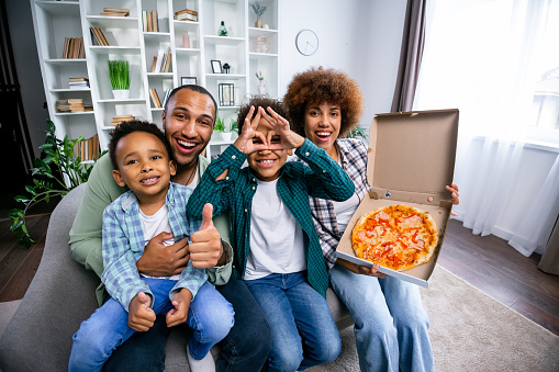 Happy Young Couple and kids Eating Pizza Sitting On sofa, Joyful African American family Enjoying Food And Looking At Camera