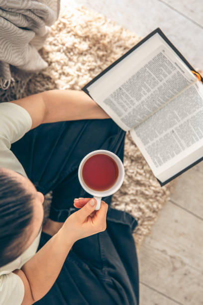 istock Top view, woman holding mug of tea while reading a book on the floor. 1830700521