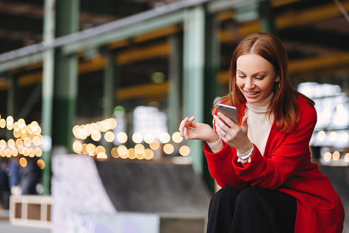 Joyful woman in red coat using smartphone with lights in background