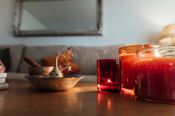 Close-up of a slow-burning incense burning in a ceramic and copper holder next to warm-colored candles. They're burning on a tabletop in a living room in front of a sofa.