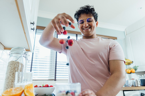 Low-angle view of a young adult dropping fresh strawberries and blueberries from a height into a plastic pot. He is wearing casual clothing and standing in his kitchen making breakfast. He is located in Durham, England.