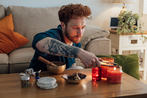 A close-up shot of a young male adult sitting indoors in front of a sofa lighting a coal pod for a diffuser next to some candles. He is having time to concentrate on well-being and mindfulness. He is located in Durham, England.