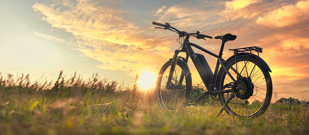 The image captures the tranquil scene of a bicycle standing in an open field at sunset. The low sun paints the sky in warm orange tones reflected in soft clouds, creating an electric picturesque backdrop. The bike is seen in profile, with emphasis on the sturdy frame, electric drivetrain and shiny spokes of the wheels. The foreground features grasses and wildflowers that appear slightly blurred in the soft evening light, adding atmosphere to the scene