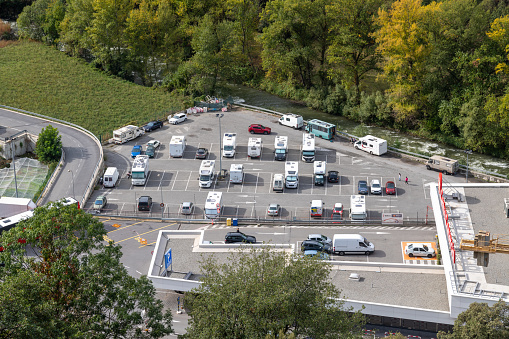 A motorhome parking space from above in Andorra in the parking lot of a shopping center. You can see some motorhomes parked on marked areas. In the background are trees and a rushing river