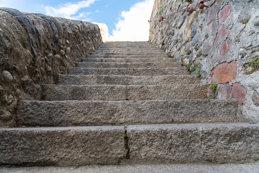 A stone staircase leading upwards, lined by a high stone wall on the right and a flat wall on the left. At the top you can see a blue sky with clouds