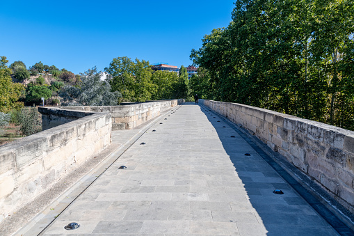 Stone footbridge from above with neatly worked stone and a stone parapet on both sides. Small spotlights are carved into the ground. To the right of the bridge are trees under a blue sky