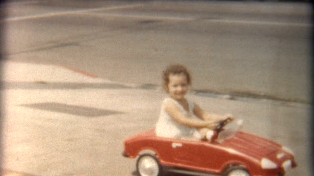 A little girl rides in her red car next to other vehicles in the 1960's. 