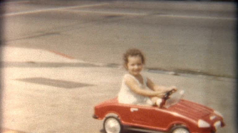 A little girl rides in her red car next to other vehicles in the 1960's.