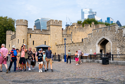 On a sunny September day, groups of tourists are walking along the Embankment near the Tower of London. Some of the buildings in the City of London can be seen above the crenellations.