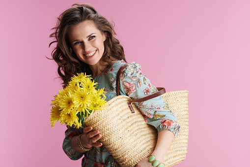 Portrait of smiling trendy female with long wavy brunette hair with yellow chrysanthemums flowers and straw bag isolated on pink background.