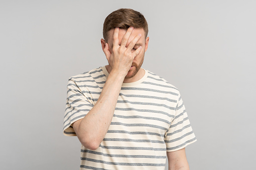 Frustrated man lowering head and eyes covers face with hand. Disappointed stressed middle aged male making facepalm gesture with hand, blaming himself for mistake isolated on studio gray background