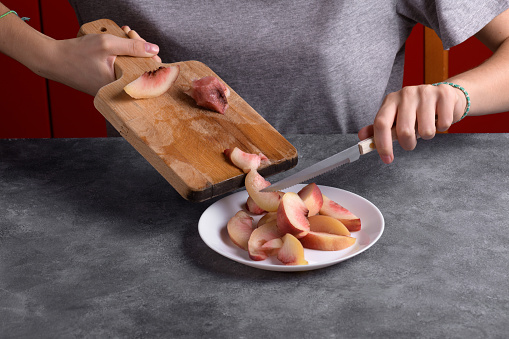 Authentic female hands cutting a fresh ripe peach on wooden cutting board on a grey table background, close up. Healthy summer snack, food lifestyle