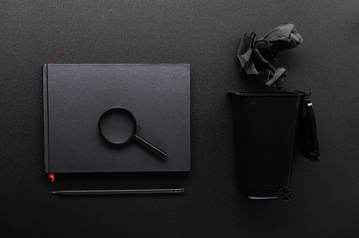 Magnifying glass on a notepad. Composition of black objects. Notebook and trash can. Search for information.