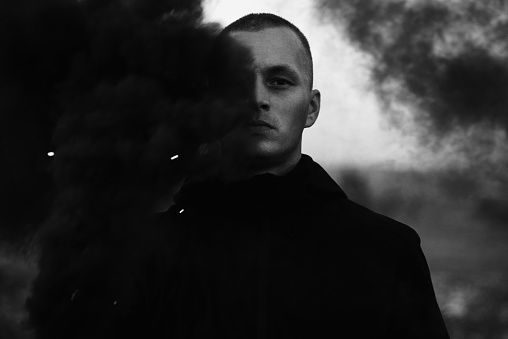 Portrait of a serious sad man in the smoke of a black smoke bomb close-up in a field. Black and white photo.