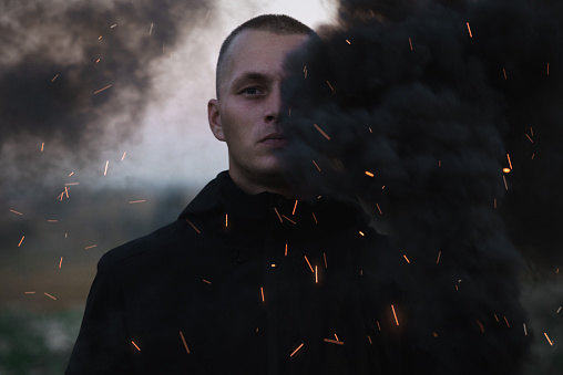 Portrait of a serious sad man in the smoke of a black smoke bomb close-up in a field.