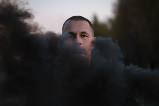 Portrait of a serious sad man in the smoke of a black smoke bomb close-up in a field.