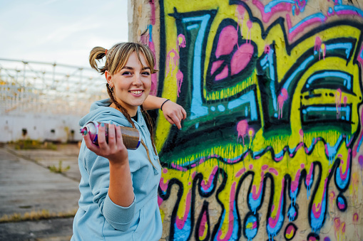 Portrait of young smiling female artist with spray can standing in front of graffiti that she made