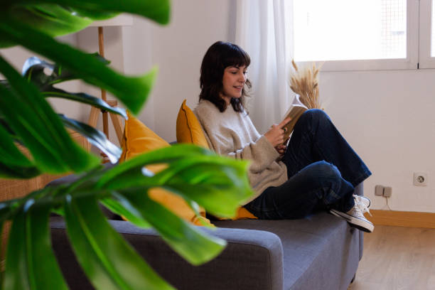 istock Young woman reading a book while sitting on a sofa at home. 1831091067
