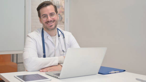 istock Middle Aged Doctor Smiling at Camera while Working on Laptop 1831105525
