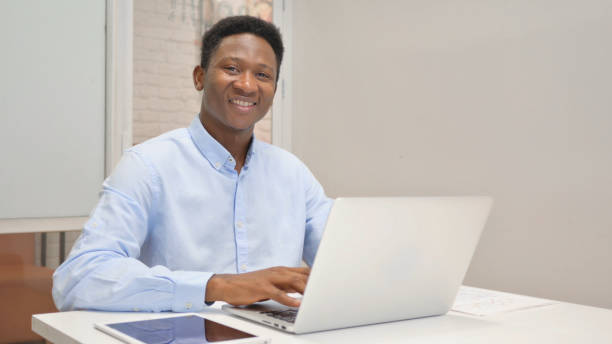 istock African Businessman Smiling at Camera while Working on Laptop in Office 1831131913