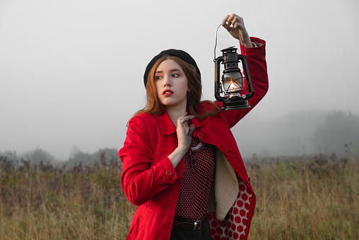 Woman with kerosene lamp in a red coat stands in a mystical misty morning field.