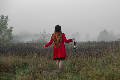 Woman with kerosene lamp in a red coat stands in a mystical misty morning field.