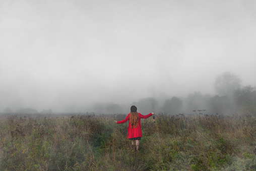 Woman with kerosene lamp in a red coat stands in a mystical misty morning field.