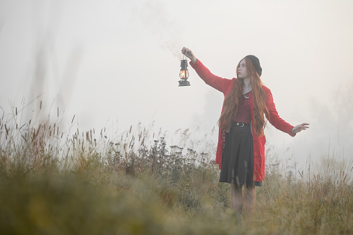 Woman with kerosene lamp in a red coat stands in a mystical misty morning field.