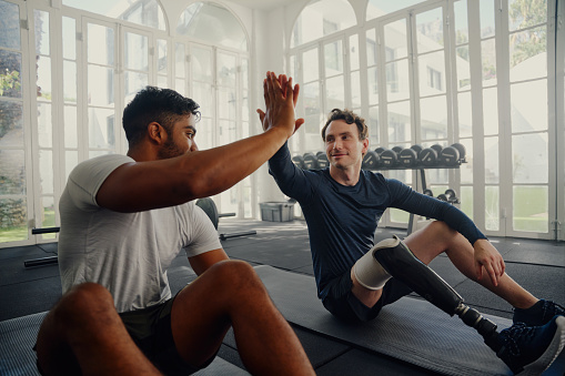 Two young multiracial men in sports clothing sitting on exercise mats and high-fiving at the gym