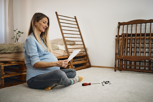 Young woman, who is expecting twins, assembling a baby crib in her bedroom.