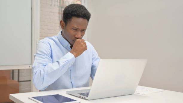 istock Coughing African Businessman Using Laptop in Office 1831140449