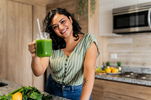Portrait of a young woman holding a glass of detox juice in kitchen at home