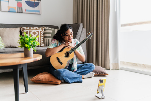 Young woman doing a live stream playing guitar in living room at house