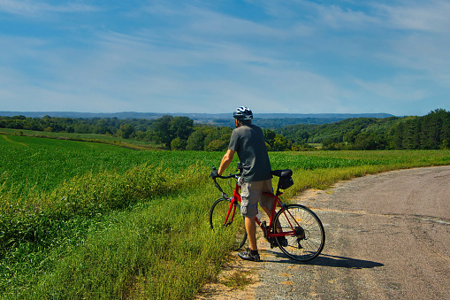 Seen from behind on a Summer day, a cyclist admires the hilltop view of fields and forests along a backroad near Arcadia, Wisconsin..