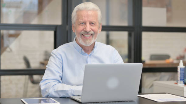 istock Old Businessman Smiling at Camera while Working on Laptop in Office 1831242226