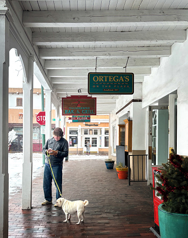 Santa Fe, NM: A senior man with a pug on the snowy Santa Fe Plaza; he’s looking down at his phone.