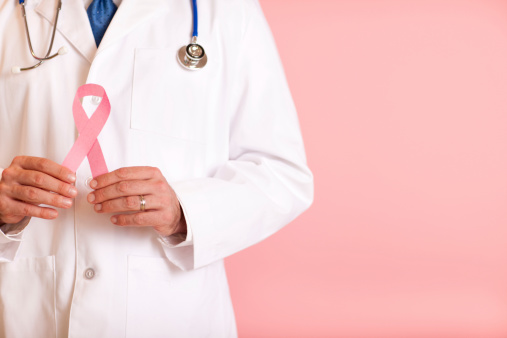 Color photo of a male doctor holding up a Breast Cancer Awareness Ribbon on pink background. Composed with room for text on right.