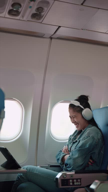 Asian female traveler enjoying a show on her tablet with headphones during the plane journey, anticipating holiday adventures.